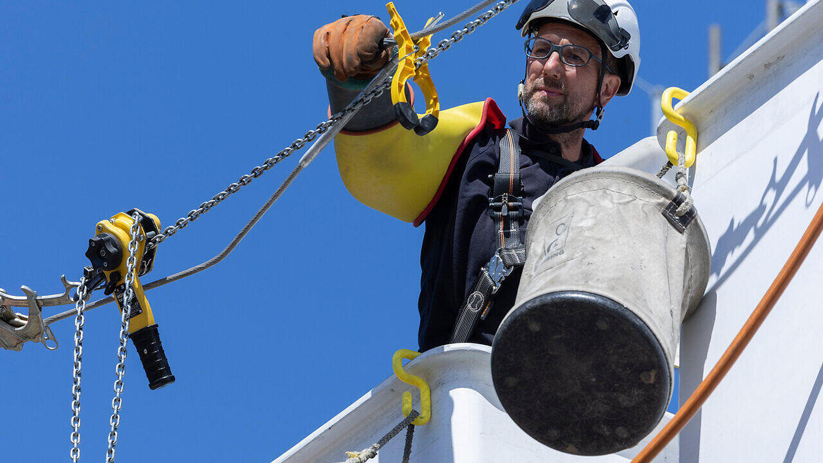 Ein Arbeiter, der mit einem Seilsystem einen Eimer an einer strukturierten Wand eines hohen Gebäudes befestigt. Der klare, blaue Himmel ist im Hintergrund sichtbar, während er mit Sicherheitsausrüstung und einem Helm ausgestattet ist.