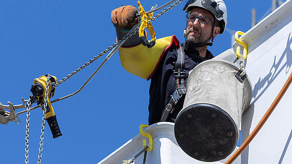 Ein Arbeiter, der mit einem Seilsystem einen Eimer an einer strukturierten Wand eines hohen Gebäudes befestigt. Der klare, blaue Himmel ist im Hintergrund sichtbar, während er mit Sicherheitsausrüstung und einem Helm ausgestattet ist.