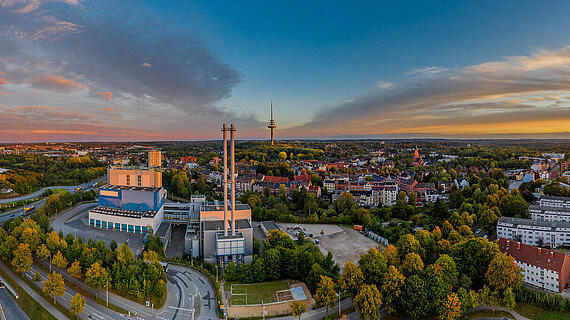 Ein Panoramablick auf eine Stadtlandschaft zum Sonnenuntergang. Im Vordergrund stehen einige Gebäude und Bäume, während im Hintergrund der Himmel in warmen Farben erleuchtet ist. Zu sehen sind auch zwei Sendemasten und die Silhouette von Wohngebäuden.