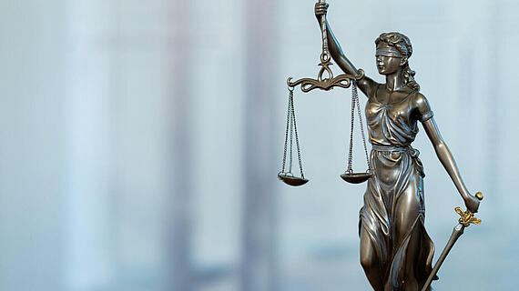 Eine bronzene Statue der Justitia, die eine Waage in der linken Hand hält und ein Schwert in der rechten Hand. Sie trägt eine Augenbinde und eine antike Robe, steht auf einem neutralen verschwommenen Hintergrund.