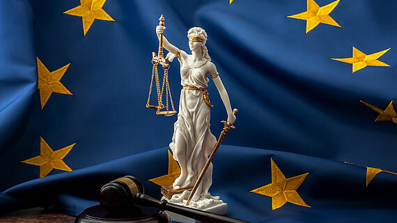 Eine Statue der Justitia mit Waage und Schwert steht vor einer blauen Flagge der Europäischen Union mit goldenen Sternen und einem Holzhammer im Vordergrund.