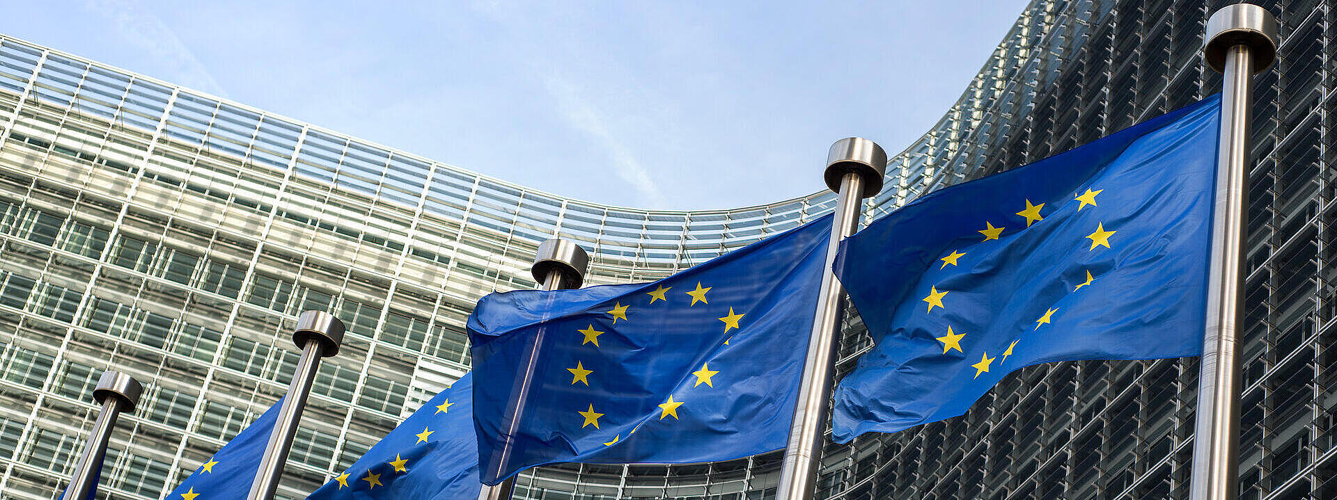 Die Flaggen der Europäischen Union wehen vor dem modernen Gebäude der Europäischen Kommission in Brüssel. Das Gebäude hat eine Glasfassade und verwandte architektonische Elemente.