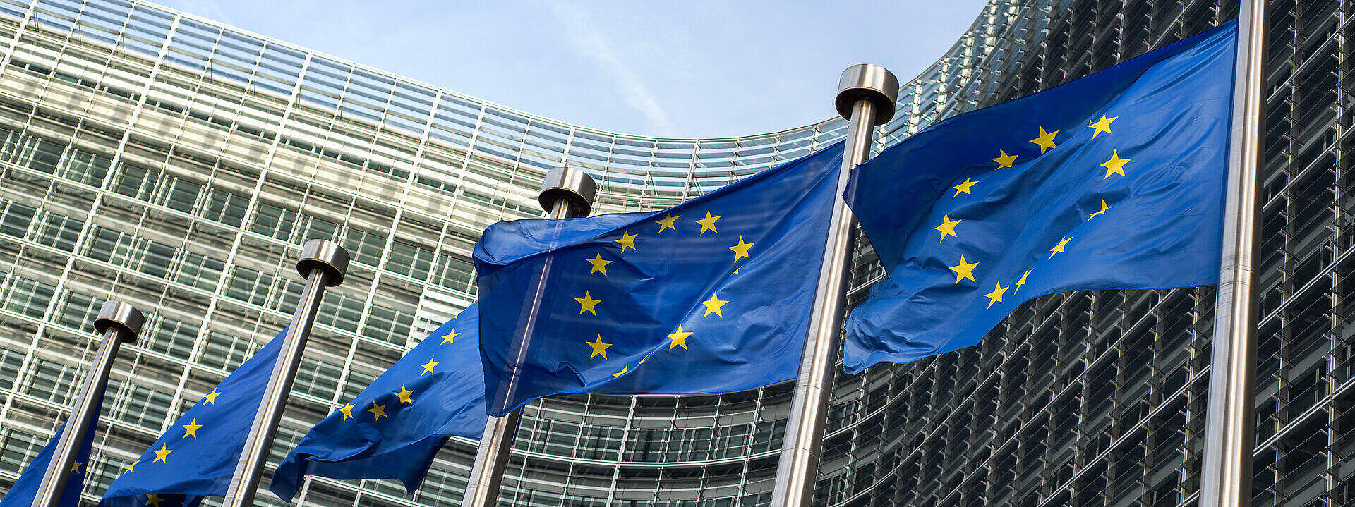 Die Flaggen der Europäischen Union wehen vor dem modernen Gebäude der Europäischen Kommission in Brüssel. Das Gebäude hat eine Glasfassade und verwandte architektonische Elemente.