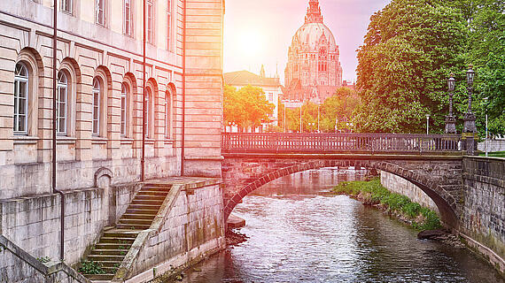 Eine Brücke über einen Fluss, umgeben von Bäumen und historischen Gebäuden. Im Hintergrund steht eine markante Kuppelarchitektur, die von Sonnenlicht beleuchtet wird.