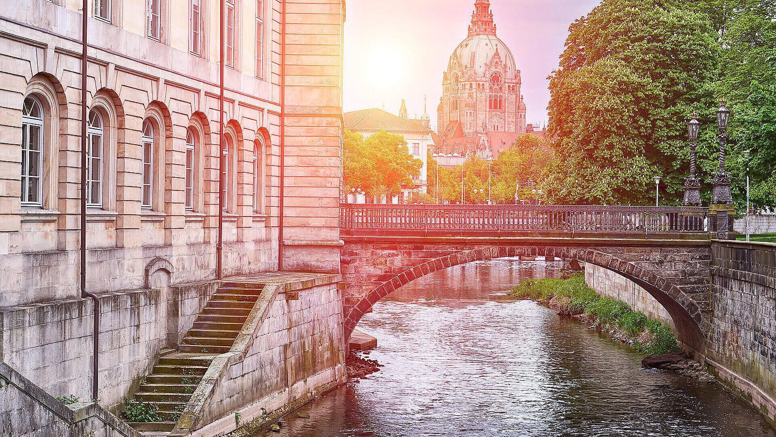 Eine Brücke über einen Fluss, umgeben von Bäumen und historischen Gebäuden. Im Hintergrund steht eine markante Kuppelarchitektur, die von Sonnenlicht beleuchtet wird.