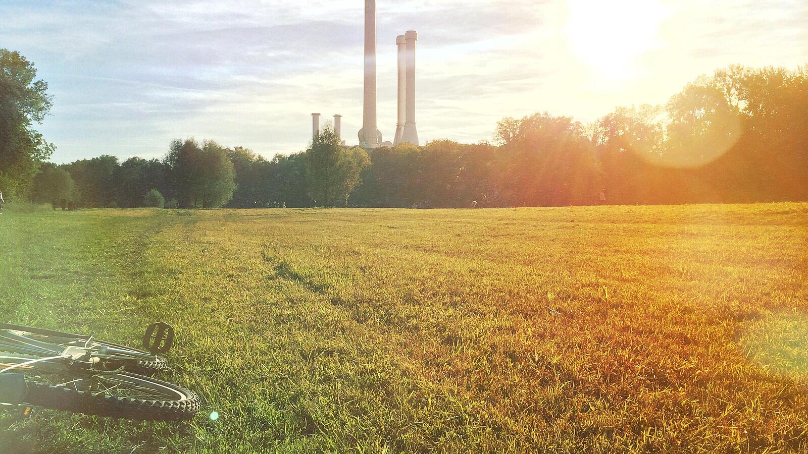 Grüne Wiese mit Fahrrad im Vordergrund, im Hintergrund Schornsteine einer Industrieanlage bei Sonnenuntergang.