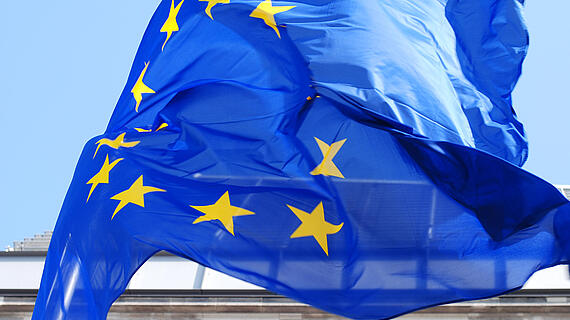 Eine wehende Flagge der Europäischen Union mit einem blauen Hintergrund und einem Kranz aus zwölf goldenen Sternen. Im Hintergrund sind Gebäudefenster sichtbar.