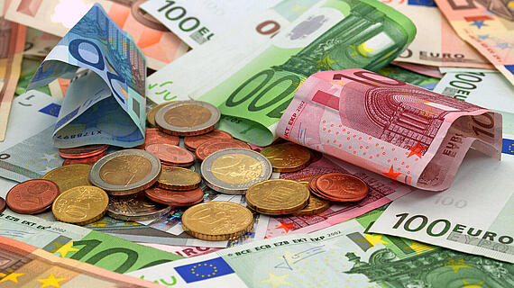 Ein Haufen Euro-Banknoten und Münzen auf einer Tischoberfläche. Zentrale sind mehrere 100- und 50-Euro-Scheine zu sehen, umgeben von verschiedenen Münzen. Eine gefaltete 5-Euro-Note liegt oberhalb der Banknoten.