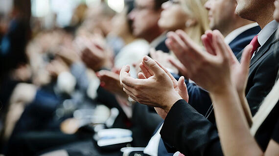Eine Gruppe von Menschen, die in einem Konferenzraum sitzt, applaudiert. Die Hände mehrerer applaudierender Zuschauer sind im Vordergrund sichtbar, während die Gesichter der Anwesenden teilweise zu sehen sind.