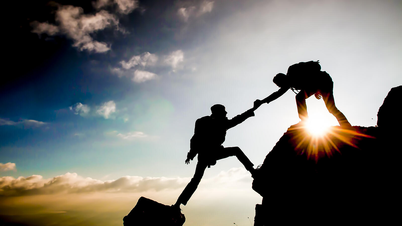 Silhouetten zweier Bergsteiger, einer hilft dem anderen beim Aufstieg im Sonnenuntergang.