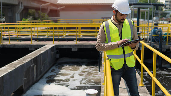 Ein Mann in einem gelben Sicherheitsanzug und einem weißen Helm steht auf einer Plattform neben einem klärenden Wasserbecken. Er hält ein Tablet in der Hand und schaut darauf, während sich im Hintergrund technische Anlagen und Gebäude befinden.