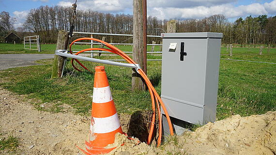 Ein grauer Sicherungskasten steht neben einem offenen Erdloch, aus dem orangefarbene Kabel herausführen. Ein orange-weißer Verkehrsleitkegel steht in der Nähe auf dem Boden. Im Hintergrund sind Bäume und ein Zaun sichtbar.
