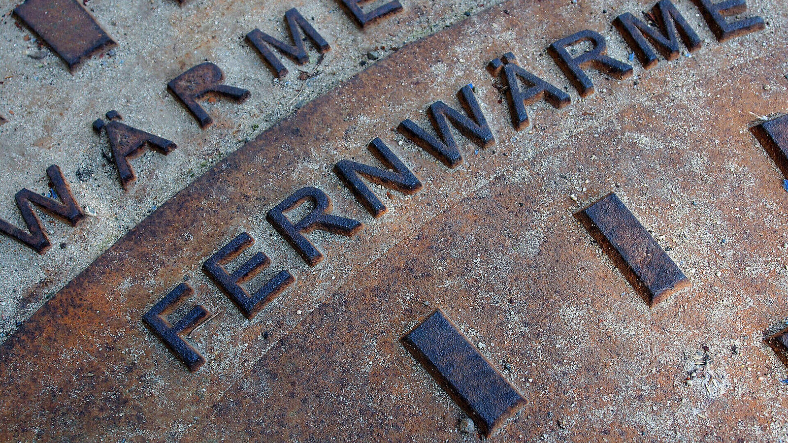 Eine Nahaufnahme von einem Gussdeckel mit der Aufschrift "FERNWÄRME" und weiteren kleinen, unleserlichen Schriftzügen, auf einer rissigen Oberfläche.