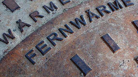 Eine Nahaufnahme von einem Gussdeckel mit der Aufschrift "FERNWÄRME" und weiteren kleinen, unleserlichen Schriftzügen, auf einer rissigen Oberfläche.