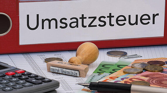 Ein Tisch mit der Aufschrift „Umsatzsteuer“, auf dem ein Taschenrechner, Münzen, Euro-Scheine und ein Stempel liegen.