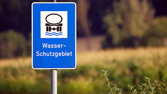 Ein blauer Schilder mit dem Symbol eines Wasserbeckens und der Aufschrift "Wasser-Schutzgebiet" steht in einer grünen Umgebung, umgeben von Feldern und Bäumen im Hintergrund.