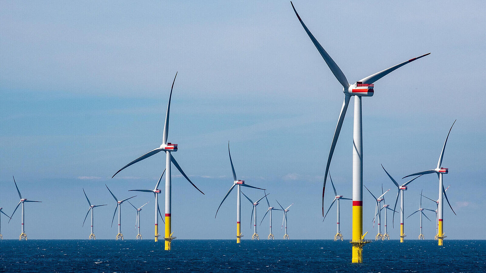 Eine Reihe von Windkraftanlagen im Meer, die auf gelben Fundamenten montiert sind. Der Himmel ist klar und blau, und die Windräder sind in verschiedenen Positionen ausgerichtet, während sie sich um einen zentralen Punkt gruppieren.