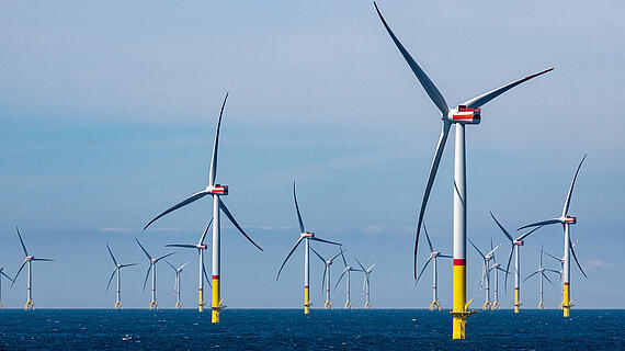 Eine Reihe von Windkraftanlagen im Meer, die auf gelben Fundamenten montiert sind. Der Himmel ist klar und blau, und die Windräder sind in verschiedenen Positionen ausgerichtet, während sie sich um einen zentralen Punkt gruppieren.