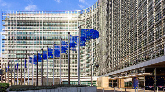 Das Gebäude der Europäischen Kommission in Brüssel ist modern gestaltet. Vor dem Gebäude stehen mehrere Fahnen mit dem EU-Logo an Fahnenmasten, während die glasverkleidete Architektur des Gebäudes im Hintergrund sichtbar ist.