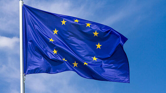 Die Flagge der Europäischen Union weht vor einem klaren, blauen Himmel. Die Flagge ist dunkelblau mit zwölf goldenen Sternen, die einen Kreis bilden, und ist am oberen Ende eines Flaggenmastes befestigt.