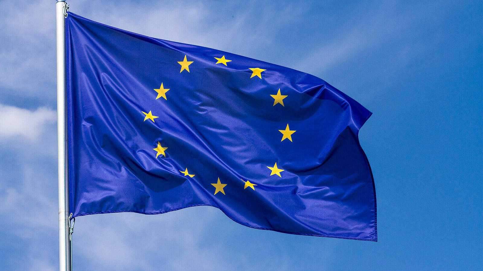 Die Flagge der Europäischen Union weht vor einem klaren, blauen Himmel. Die Flagge ist dunkelblau mit zwölf goldenen Sternen, die einen Kreis bilden, und ist am oberen Ende eines Flaggenmastes befestigt.