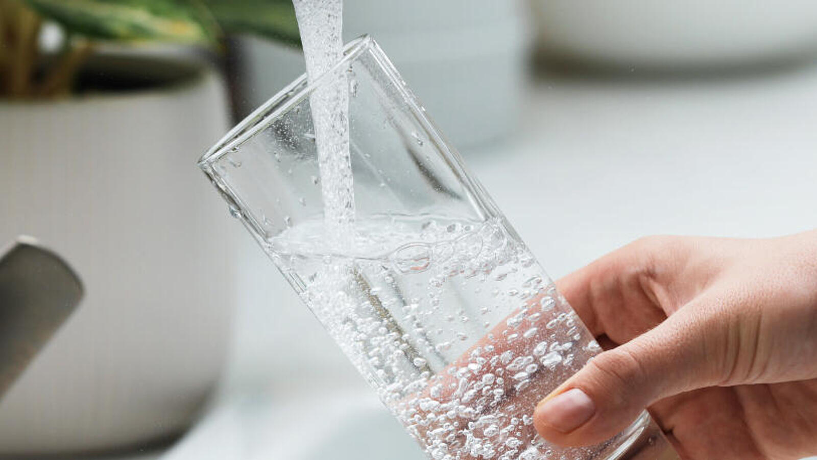 Eine Frau gießt sich an einem Haushaltswasserhahn Wasser in ihr Glas, um daraus zu trinken.