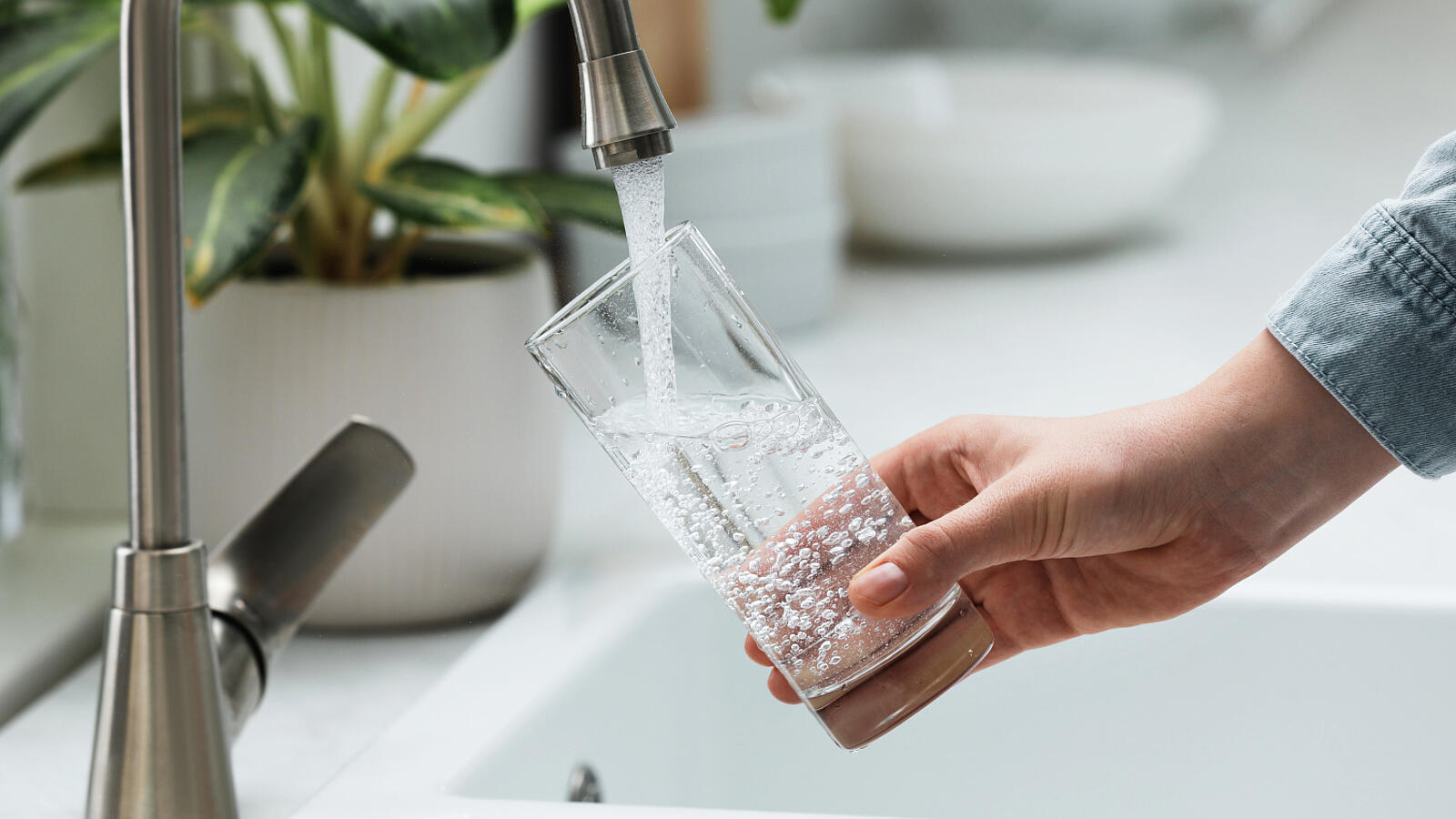 Eine Frau gießt sich an einem Haushaltswasserhahn Wasser in ihr Glas, um daraus zu trinken.