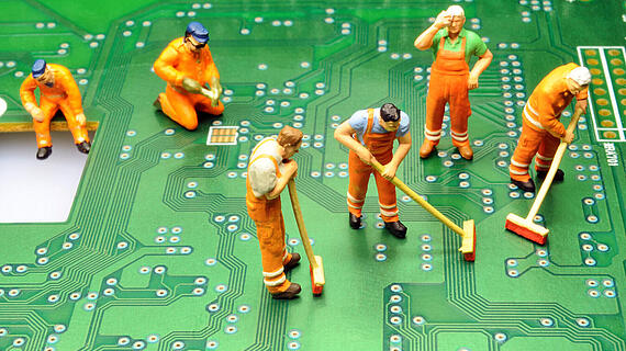 Miniaturfiguren von Arbeitern in orangefarbenen Overalls, die auf einer Leiterplatte mit grüner Farbe und elektronischen Schaltkreisen arbeiten. Einige fegen den Boden, während andere in verschiedenen Positionen stehen.