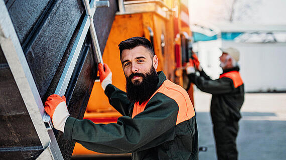 Männer in Arbeitskleidung, einer steht neben einem großen Lkw und schaut in die Kamera, der andere arbeitet im Hintergrund. Beide tragen Handschuhe und sind in einer industrialisierten Umgebung.