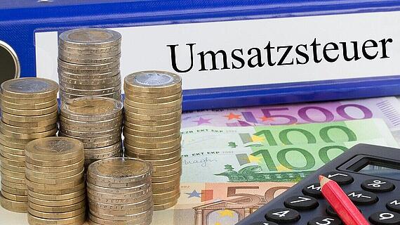 Ein Stapel von Münzen, der neben einem offenen Ordner mit der Aufschrift "Umsatzsteuer" steht. Im Hintergrund sind verschiedene Euro-Scheine, darunter 100 und 500 Euro, sowie ein Taschenrechner mit einem roten Stift zu sehen.