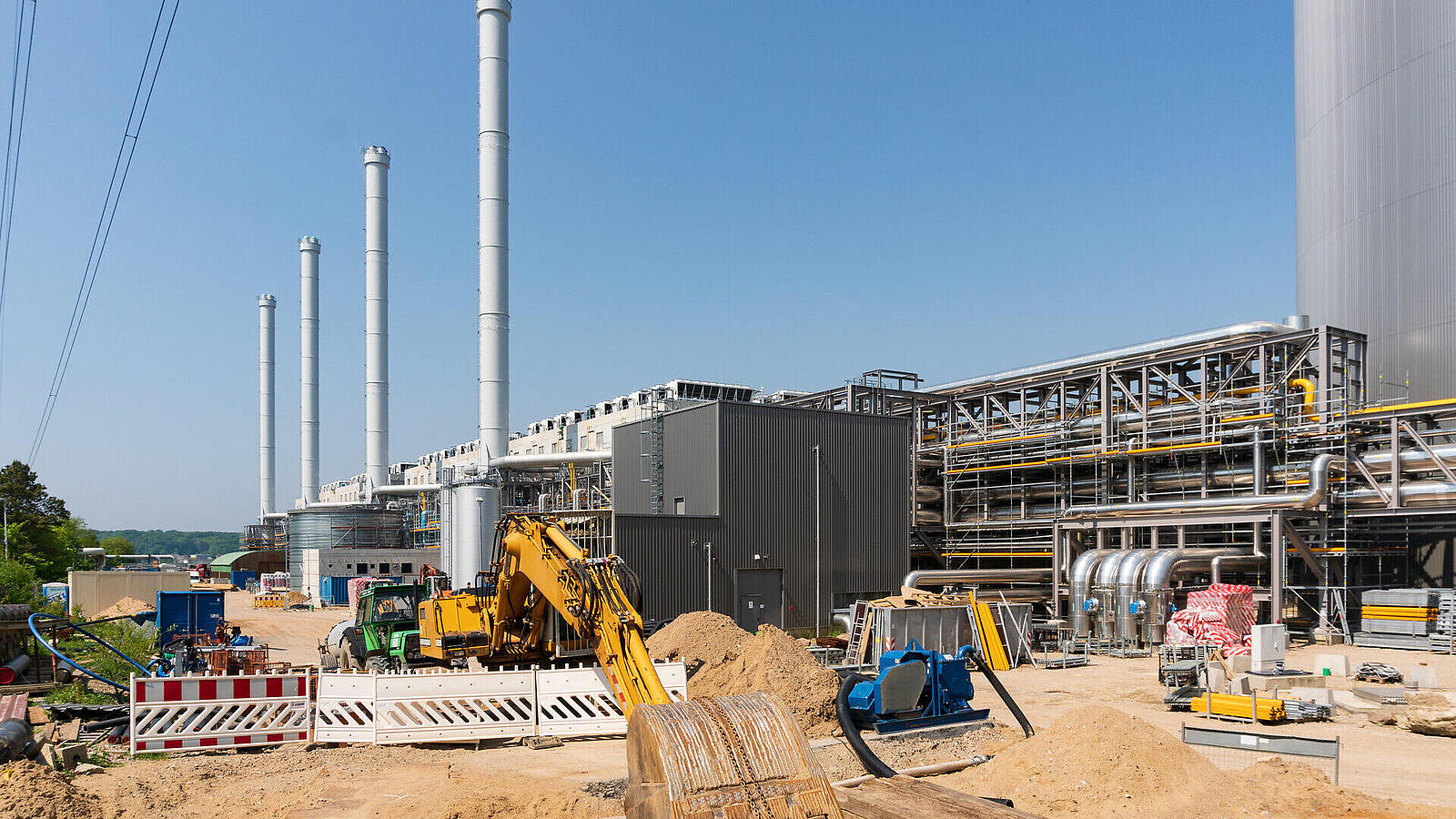 Eine Industrieanlage unter blauem Himmel, mit mehreren hohen Schornsteinen. Im Vordergrund sind Baugeräte und Baustellenmaterial zu sehen. Die Anlage besteht aus großen, metallischen Strukturen und Rohren. Sande und Erdmassen liegen im Baustellenbereich.