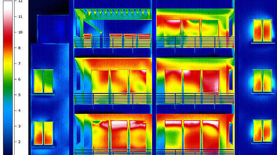 Thermografisches Bild eines Wohngebäudes mit Balkonen, das Temperaturunterschiede zeigt. Warme Bereiche erscheinen rot und gelb, kühle Bereiche in Blau und Grün. Eine Skala links zeigt die Temperatur in Grad Celsius an.