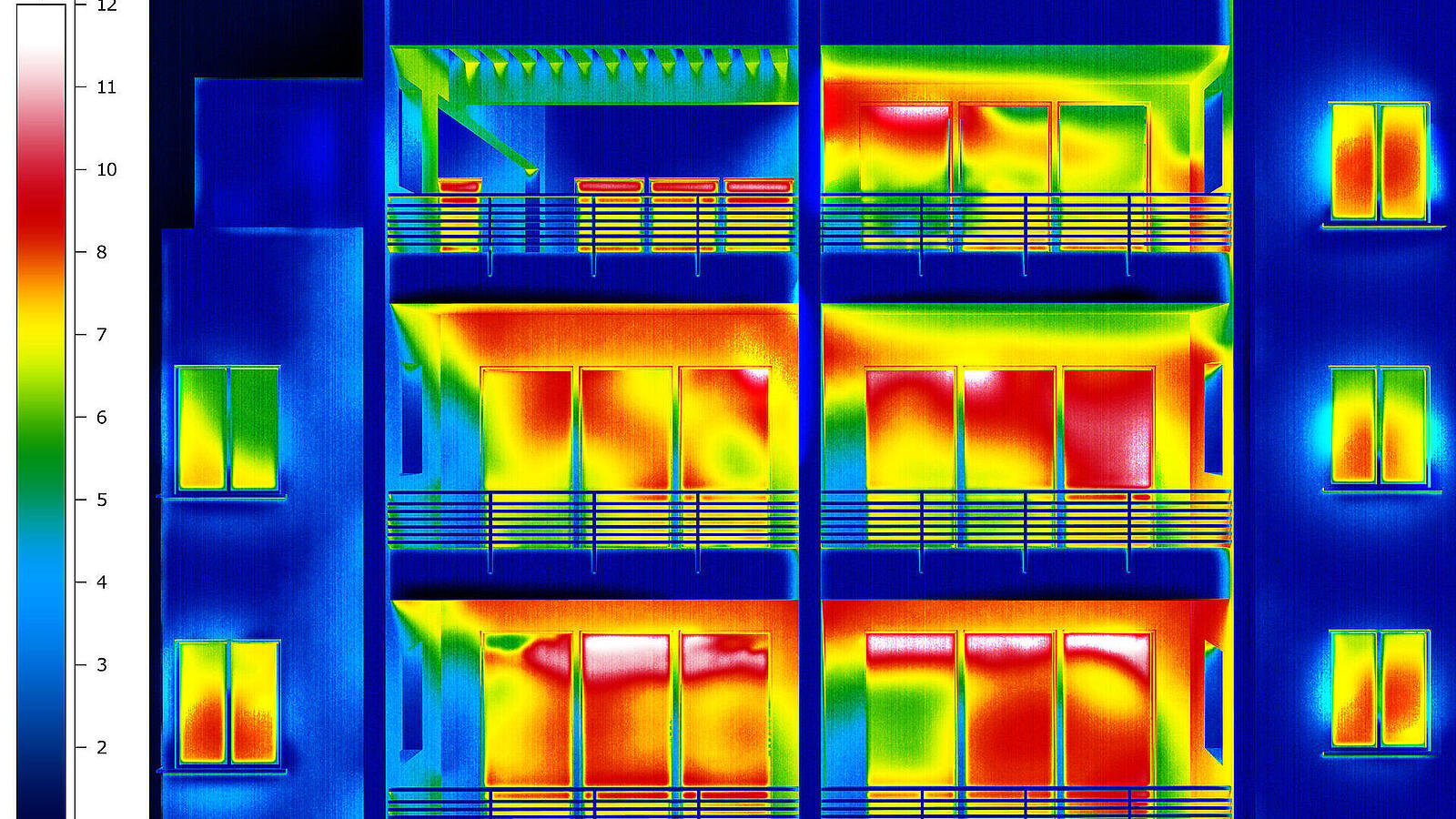 Thermografisches Bild eines Wohngebäudes mit Balkonen, das Temperaturunterschiede zeigt. Warme Bereiche erscheinen rot und gelb, kühle Bereiche in Blau und Grün. Eine Skala links zeigt die Temperatur in Grad Celsius an.