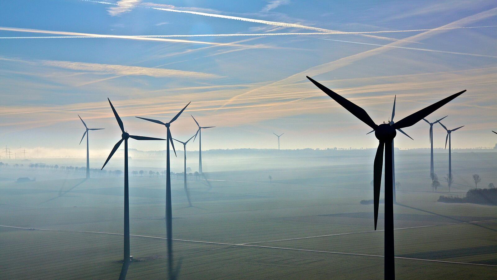 Windräder stehen in einer nebligen Landschaft unter blauem Himmel mit Kondensstreifen.