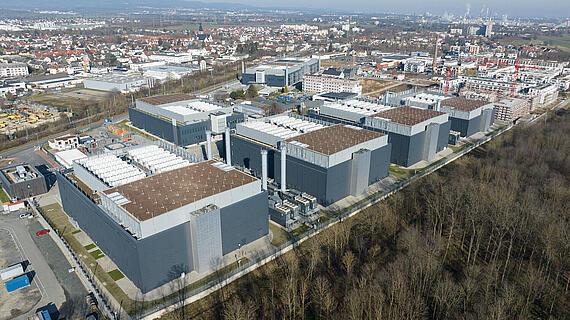 Luftaufnahme eines Industriegebiets mit mehreren großen, grauen Gebäuden. Im Vordergrund sind Lagerflächen und Parkplätze sichtbar, während im Hintergrund Wohngebiete und eine städtische Landschaft zu sehen sind. Der Himmel ist klar und die Landschaft erstreckt sich bis zu den Bergen.