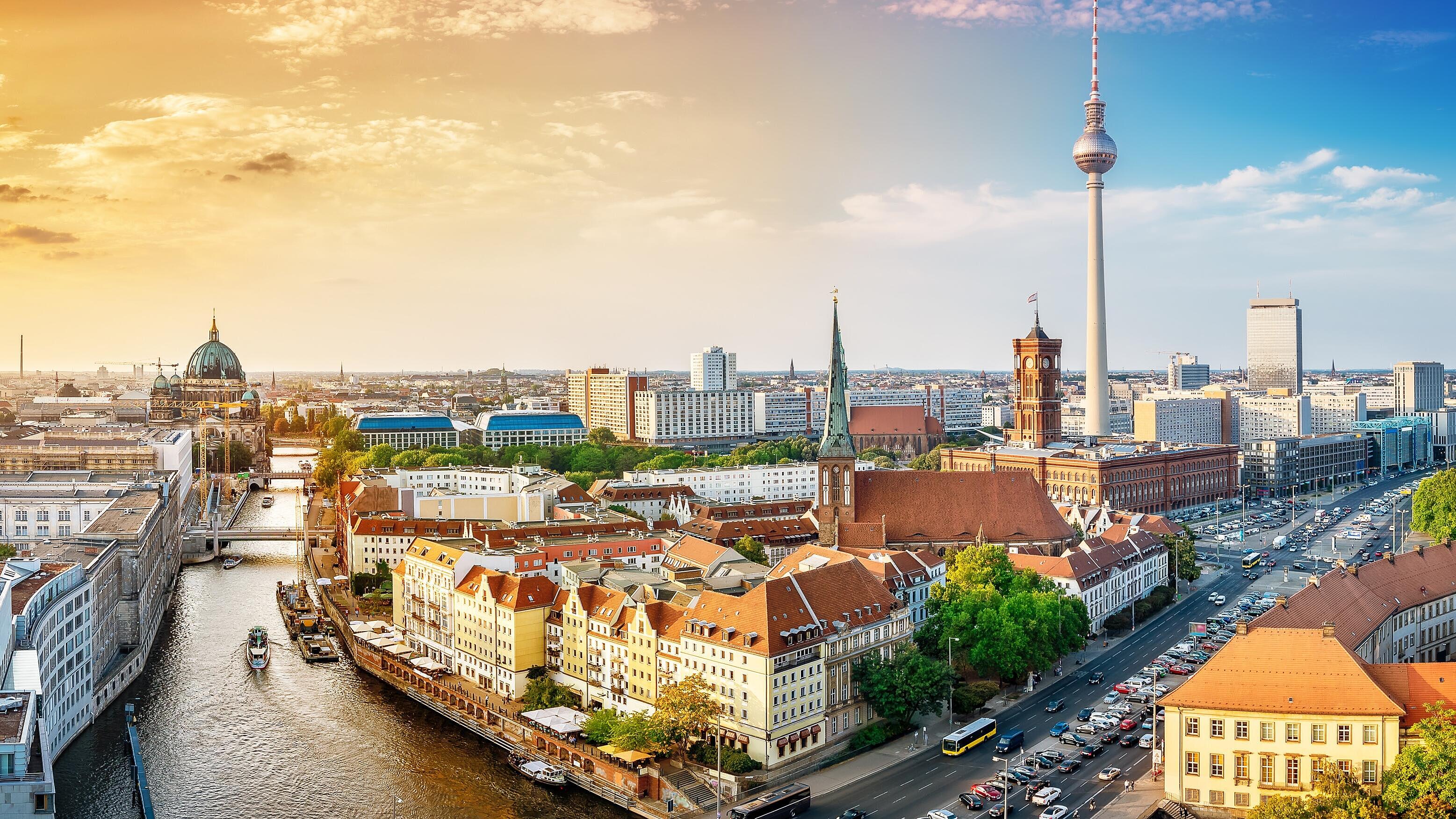 Panoramablick auf Berlin während des Sonnenuntergangs. Die Stadt zeigt eine Mischung aus modernen und historischen Gebäuden, darunter der Berliner Fernsehturm. Ein Fluss fließt durch die Stadt, umgeben von Bäumen und Straßen mit Fahrzeugen.