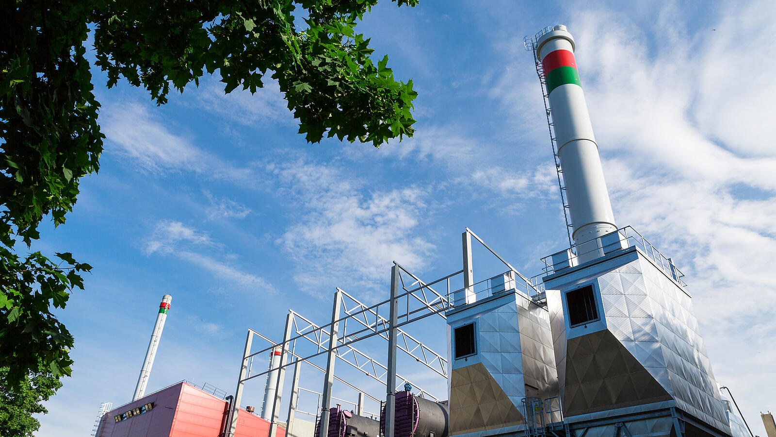 Industrieanlage mit hohem Schornstein und metallischen Gebäudestrukturen unter blauem Himmel mit Wolken.