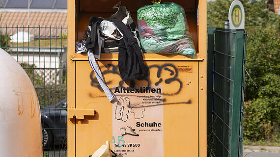 Ein überfüllter Altkleidercontainer mit Müllsäcken darunter. Auf dem Container steht "Altkleider Schuhe". Im Hintergrund sind Bäume und ein Straßenschild zu sehen.