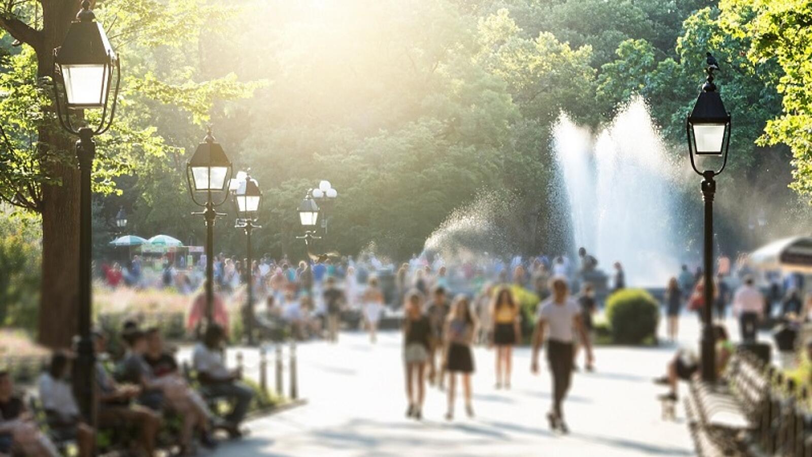 Menschen spazieren durch einen sonnigen Park mit Springbrunnen.