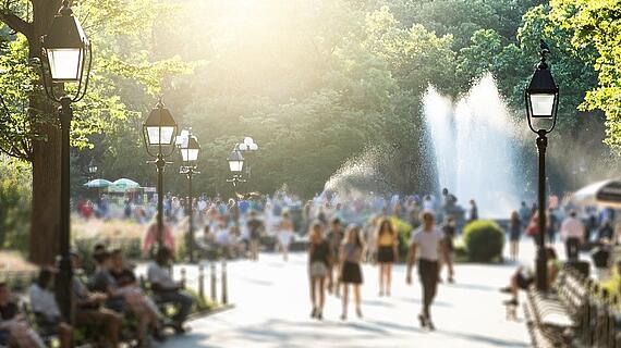 Menschen spazieren durch einen sonnigen Park mit Springbrunnen.
