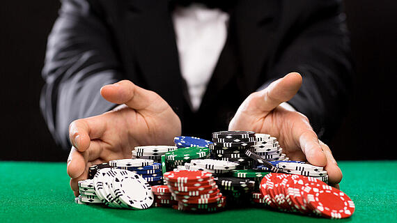 Ein Mann in einem Smoking streckt seine Hände aus, um eine Gruppe von Pokerchips zu präsentieren. Die Chips sind in verschiedenen Farben, darunter Schwarz, Weiß, Rot und Grün, und liegen auf einem grünen Spieltisch.