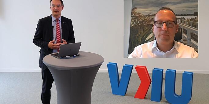 Ein Mann steht an einem Stehtisch mit einem Laptop vor sich. Neben ihm stehen die Buchstaben 'VKU'. Im rechten oberen Eck ist ein zweites Bild eines Mannes eingeblendet.