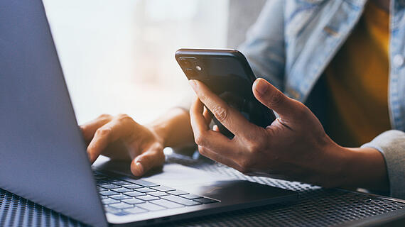 Eine Hand, die ein Smartphone hält, während die andere Hand auf einer Laptop-Tastatur tippt. Der Laptop steht auf einem Netzuntergrund, und die Person trägt eine Jeansjacke und ein gelbes Oberteil.