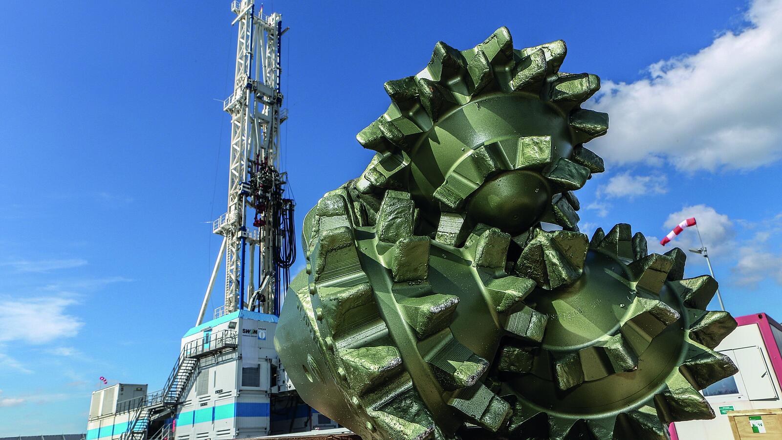 Großer metallischer Bohrkopf mit mehreren gezackten Fräskegeln liegt im Vordergrund auf einer Holzfläche. Im Hintergrund steht ein hoher Bohrturm mit weiß-blauen Streifen, unter klarem Himmel mit wenigen Wolken. Rechts ist ein Windrichtungsanzeiger sichtbar.