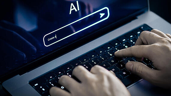 Eine Person tippt auf einer Laptop-Tastatur. Auf dem Bildschirm sind die Wörter 'AI' und ein Texteingabefeld mit der Aufforderung 'Create' zu sehen. Der Hintergrund ist leicht unscharf und dunkel.