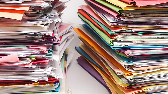 Zwei hohe Stapel von Dokumenten, einer links mit buntem Papier und einer rechts mit mehrfarbigem, unordentlichem Papier, stehen nebeneinander auf einer hellen Oberfläche.