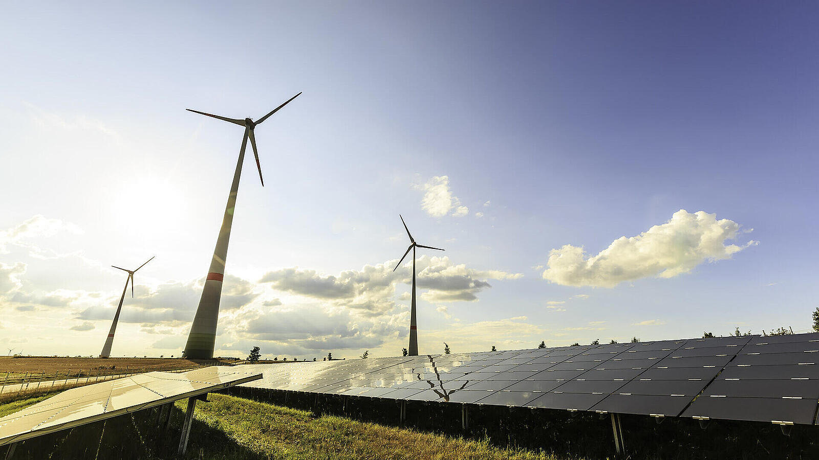 Windkraftanlagen neben einer Solaranlage in einer offenen Landschaft unter einem klaren Himmel mit einigen Wolken.