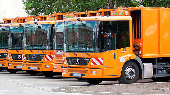 Fünf orange Müllfahrzeuge, die nebeneinander auf einem Parkplatz stehen. Jedes Fahrzeug trägt Sicherheitsstreifen und hat eine Werbebotschaft auf der Seite.