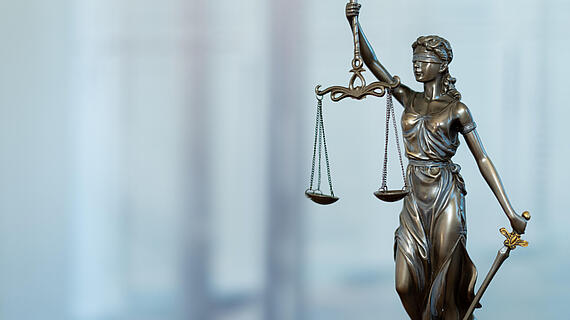 Eine bronzene Statue der Justitia, die eine Waage in der linken Hand hält und ein Schwert in der rechten Hand. Sie trägt eine Augenbinde und eine antike Robe, steht auf einem neutralen verschwommenen Hintergrund.