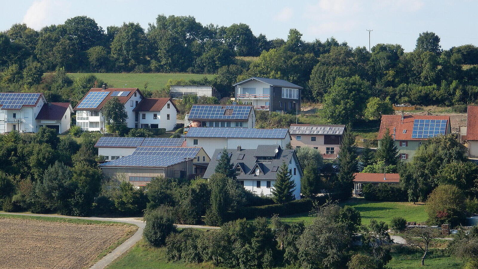 Landschaft mit mehreren Wohnhäusern, die Solarpanels auf den Dächern installiert haben. Im Hintergrund sind Bäume und ein hügeliges Feld zu sehen, unter einem klaren Himmel.